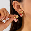Gold bow earrings with red and white checkered heart charms worn on ear